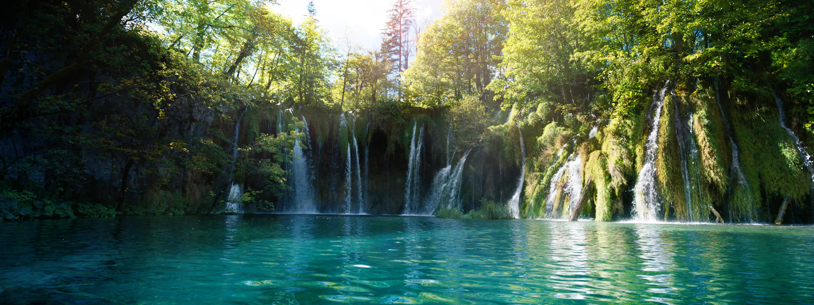 Clean Water A tranquil waterfall cascades into a clear turquoise lake, surrounded by lush green trees, under a bright, sunny sky.