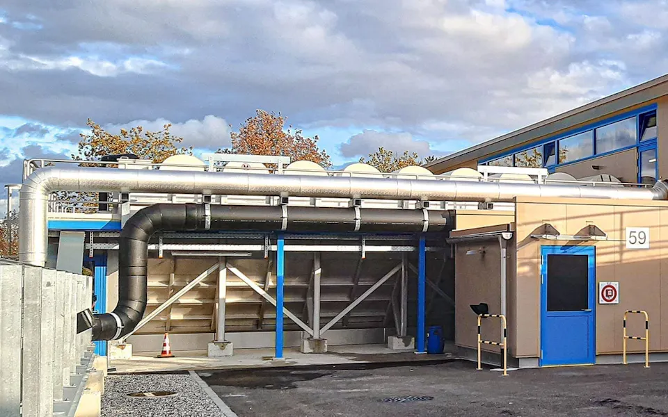 Outdoor facilities at ARA Rhein Die moderne industrielle Anlage zeigt ein beige-graues Gebäude mit mehreren Lüftungsrohren und blauer Tür, umgeben von einem gepflasterten Bereich unter einem bewölkten Himmel.