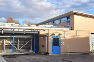 Outdoor facilities at ARA Rhein The modern industrial facility shows a beige-gray building with several ventilation pipes and a blue door, surrounded by a paved area under a cloudy sky.