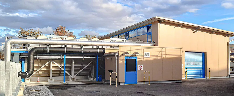 Outdoor facilities at ARA Rhein The modern industrial facility shows a beige-gray building with several ventilation pipes and a blue door, surrounded by a paved area under a cloudy sky.