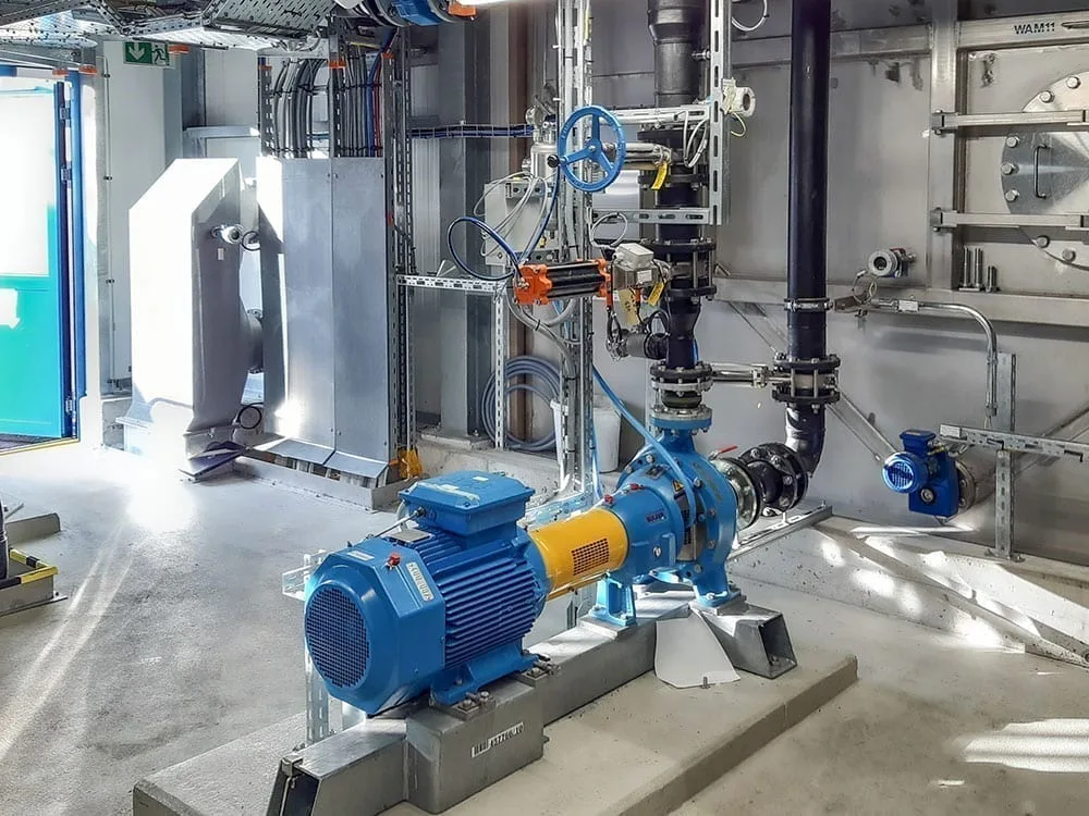 ARA Rhein Reference 3 A bright industrial room with a large blue water pump surrounded by metal pipes, control valves, and electrical wiring on a concrete floor.