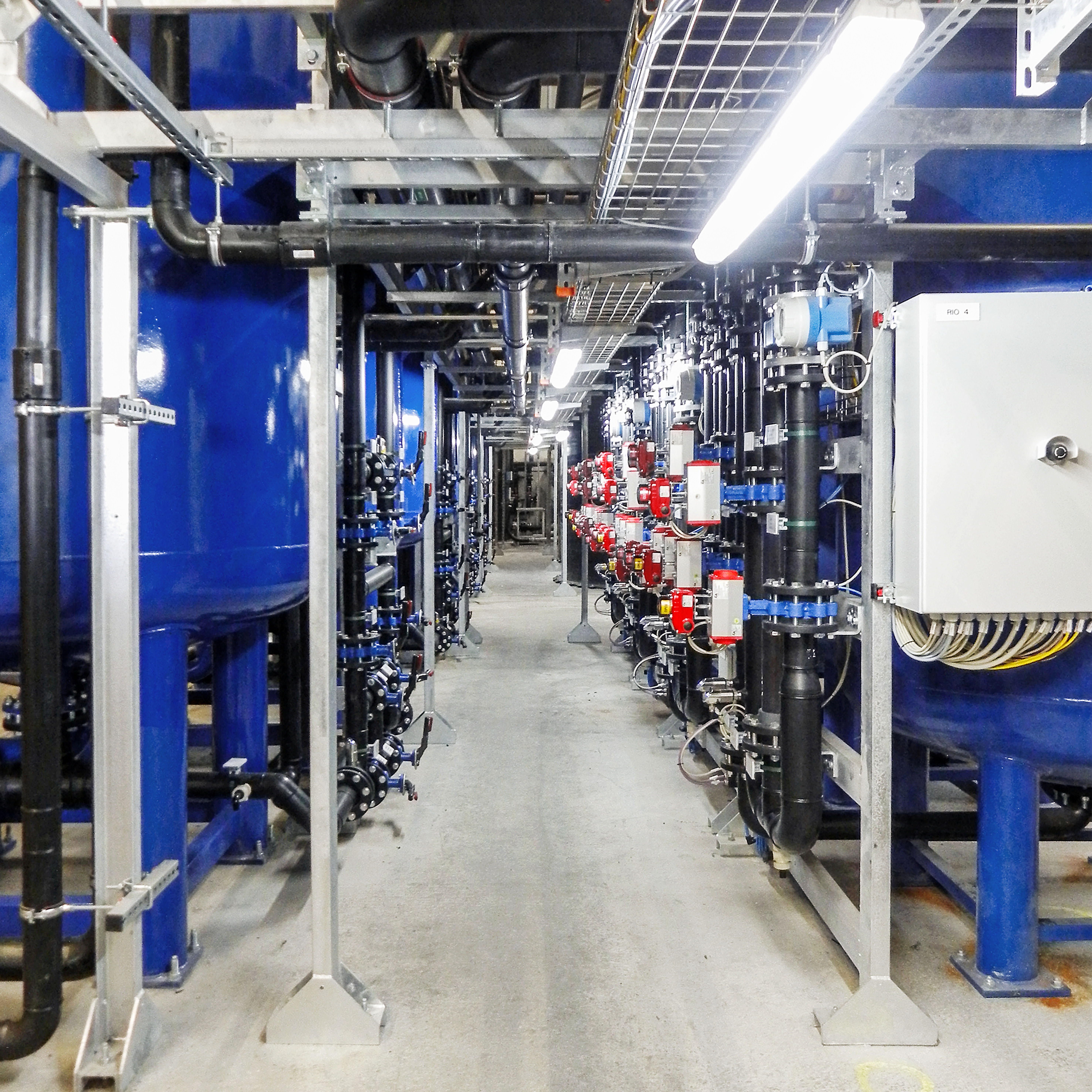 Ein industrieller Korridor mit großen blauen Tanks und komplexen Rohrleitungssystemen in einem sauber beleuchteten, technischen Umfeld.