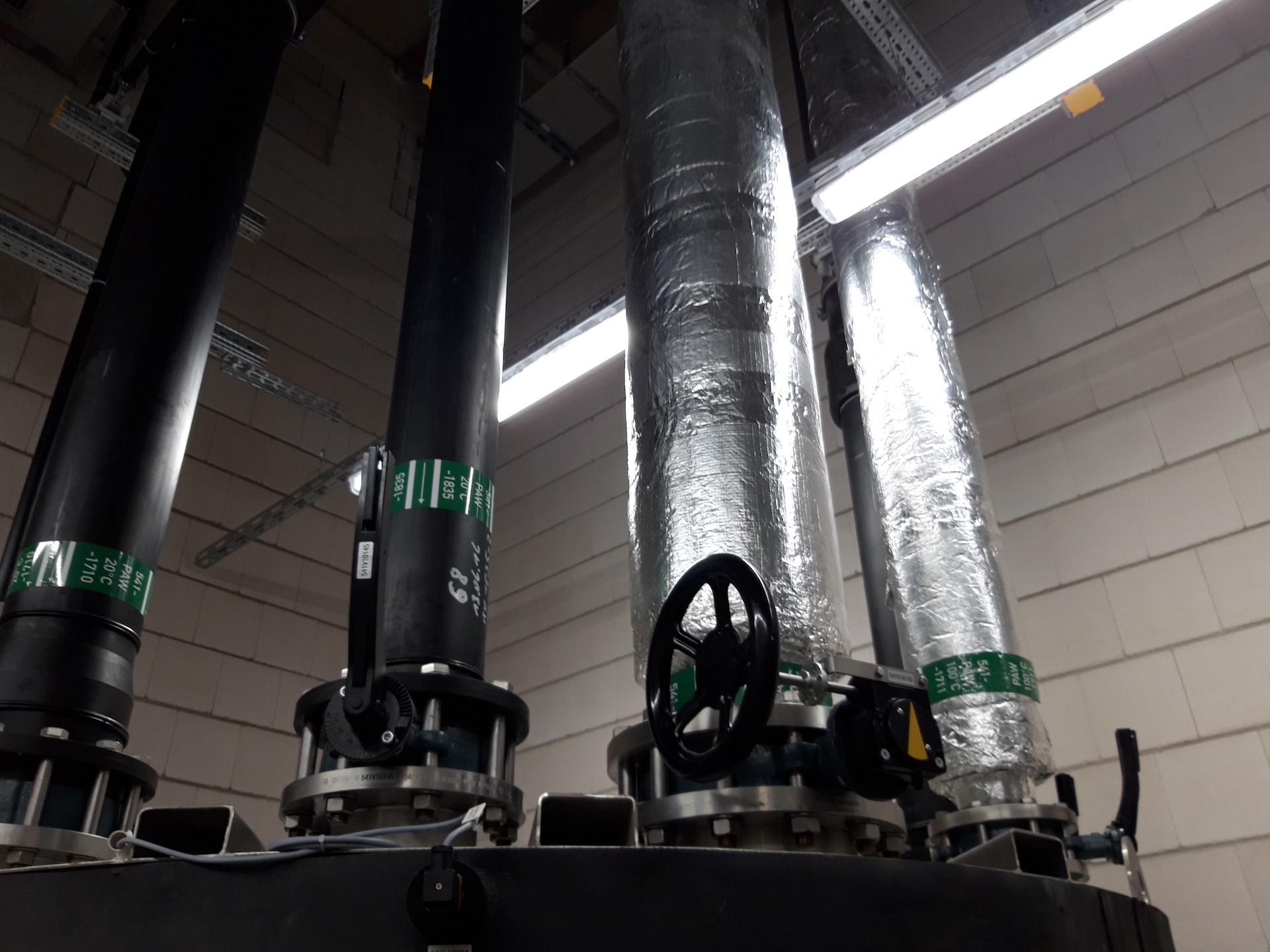 Vier große industrielle Rohre in einem technischen Raum, mit zwei isolierten silbernen Rohren und einem schwarzen Rohr mit Ventil, umgeben von neutralen grauen Wänden und fluoreszierendem Licht, verdeutlichen das Thema "Industrielle Infrastruktur und Wartung".