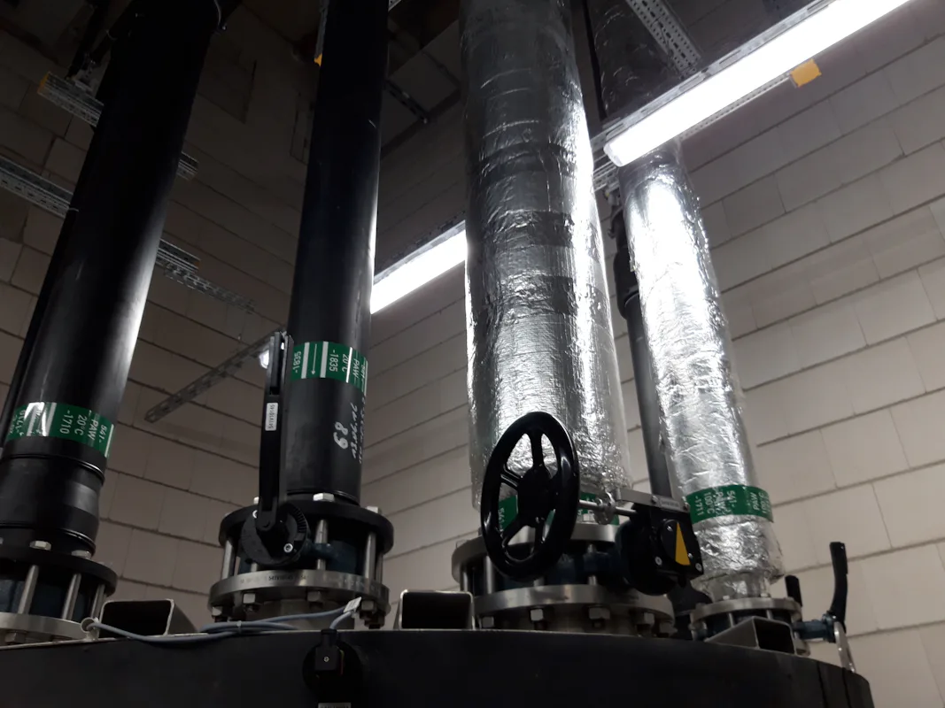 Biotest Reference Vier große industrielle Rohre in einem technischen Raum, mit zwei isolierten silbernen Rohren und einem schwarzen Rohr mit Ventil, umgeben von neutralen grauen Wänden und fluoreszierendem Licht, verdeutlichen das Thema "Industrielle Infrastruktur und Wartung".