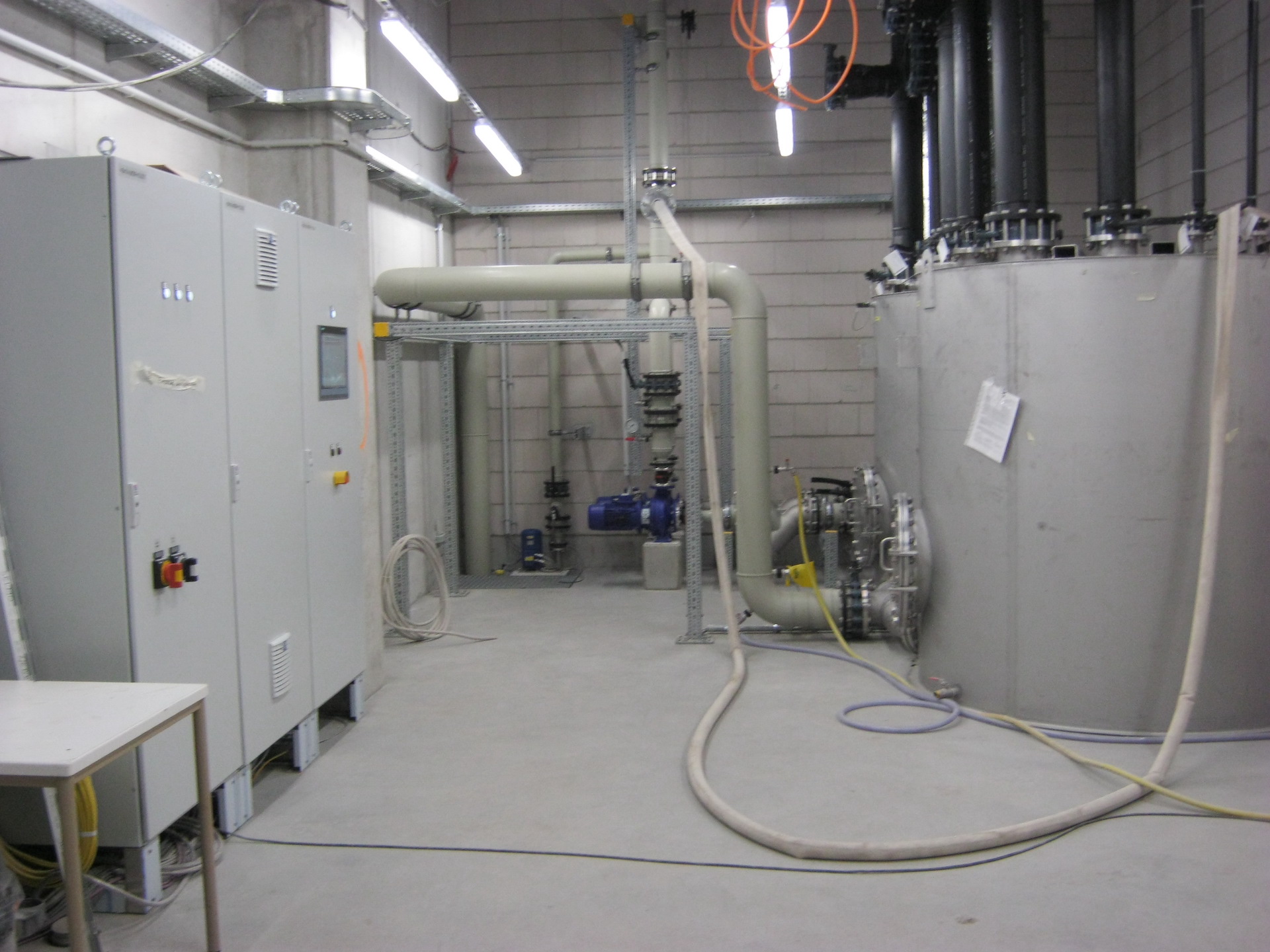 Ein moderner technischer Raum mit großen silbernen Maschinen, weißen Rohrleitungen, einer Grauschalttafel und freiliegenden Kabeln an der Decke, umgeben von Betonwänden.