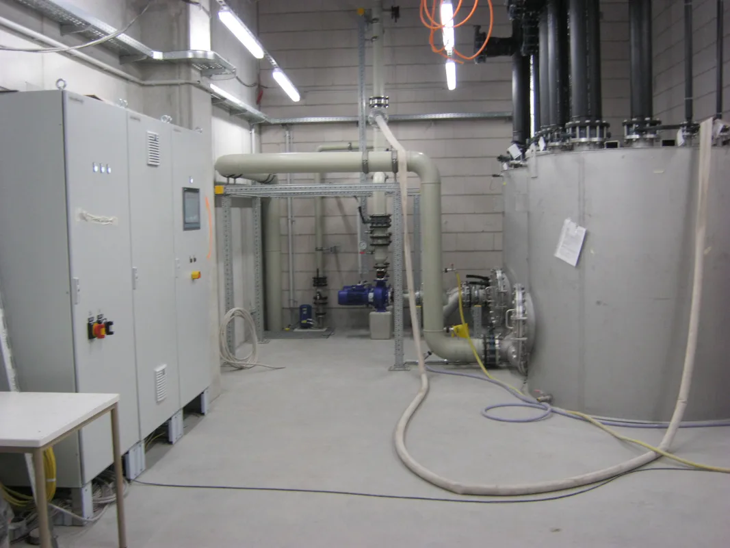 Biotest Reference Ein moderner technischer Raum mit großen silbernen Maschinen, weißen Rohrleitungen, einer Grauschalttafel und freiliegenden Kabeln an der Decke, umgeben von Betonwänden.