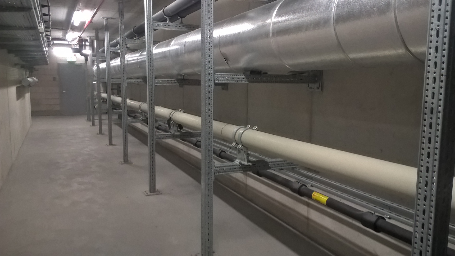 Ein grauer, industriell wirkender Technikraum mit großen, silbernen Lüftungsrohren, die entlang der Betondecke und der Wände verlaufen.