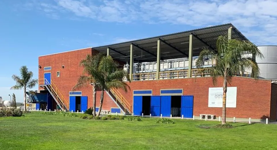 這張圖片展示了一座現代化的磚瓦建築，其特點是藍色金屬門和結構框架，前景是一片綠色草地和幾棵棕櫚樹，天空呈現晴朗的藍色。