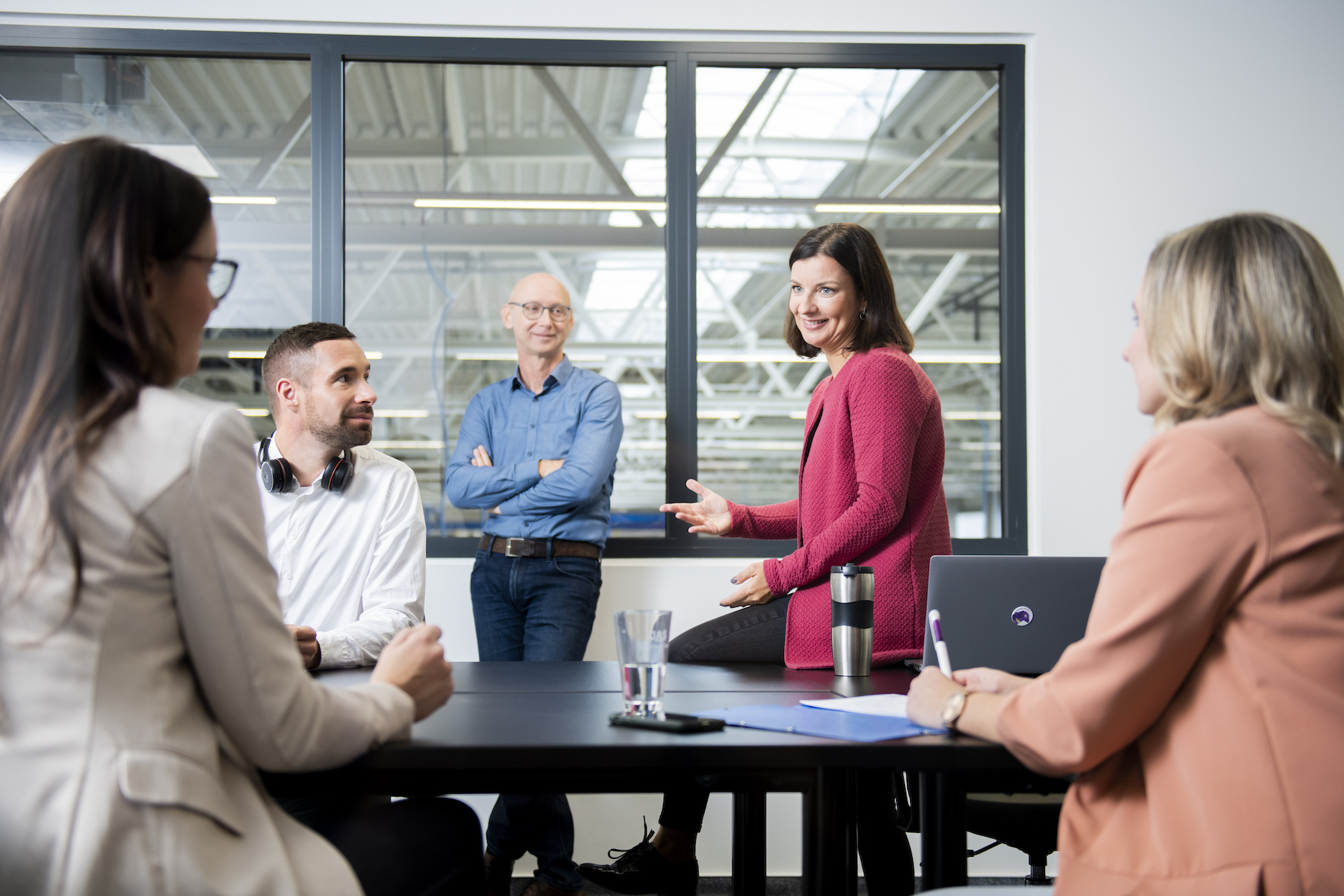 A diverse team in a modern office setting is engaged in a lively discussion around a table, with a window showing an industrial-style ceiling in the background.