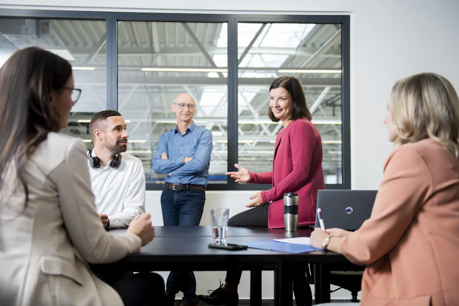 DAS Management-Team A diverse team in a modern office setting is engaged in a lively discussion around a table, with a window showing an industrial-style ceiling in the background.