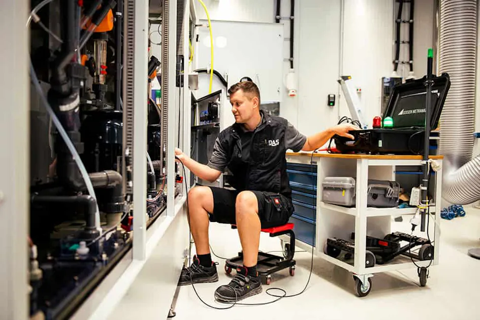 Service A technician in a workshop setting inspects large industrial machinery, seated on a rolling stool, surrounded by various tools and equipment, indicating maintenance or repair work in progress.