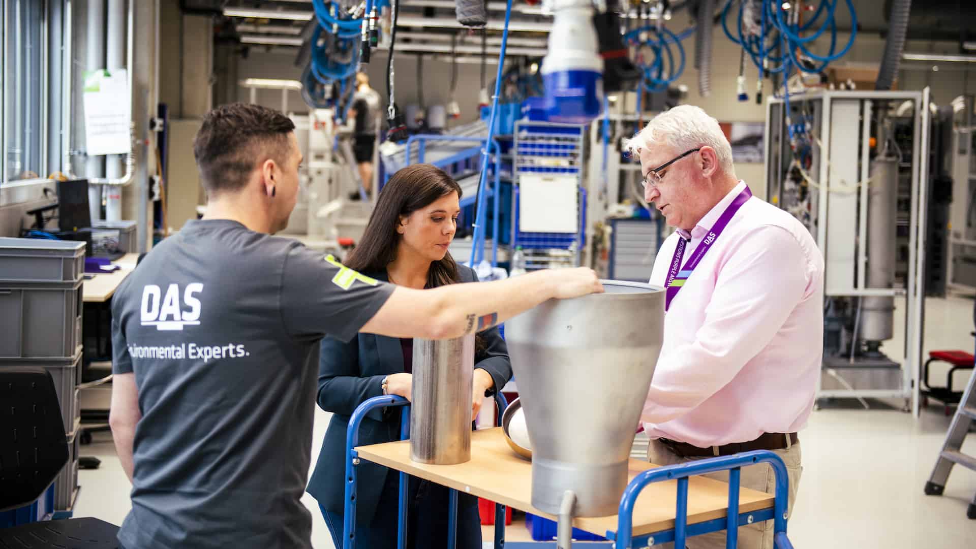 Three individuals are gathered around a table in a modern industrial workspace, examining large metallic equipment, with various tools and machinery visible in the background.
