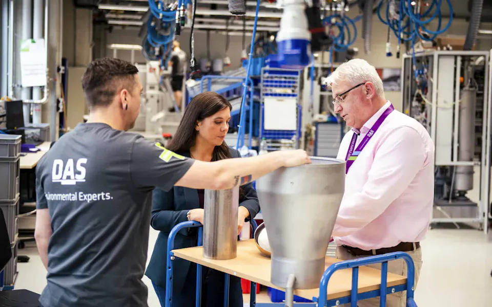DAS business unit waste gas treatment Three individuals are gathered around a table in a modern industrial workspace, examining large metallic equipment, with various tools and machinery visible in the background.