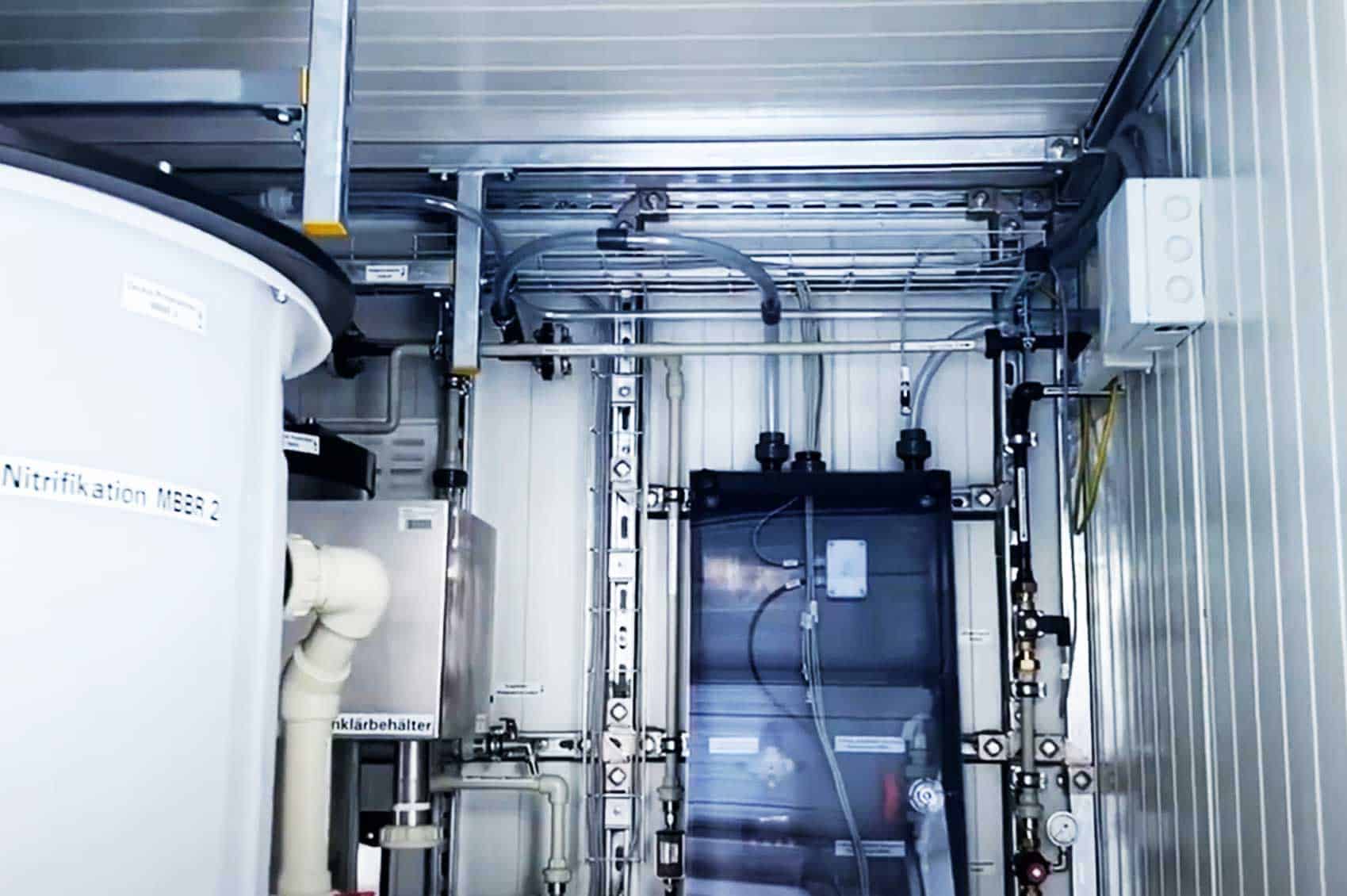 A modern industrial container with a multitude of pipes and technical devices attached to the walls, as well as a storage tank and several valves, focused on water treatment or ventilation systems.