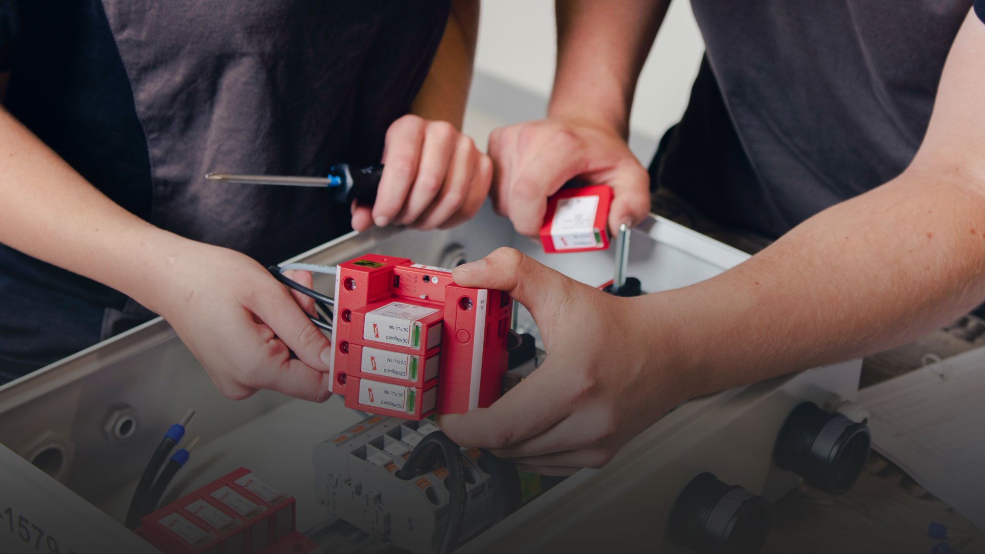 Two individuals are assembling a circuit board using red fuse blocks, while holding screwdrivers and working collaboratively inside a workshop setting.