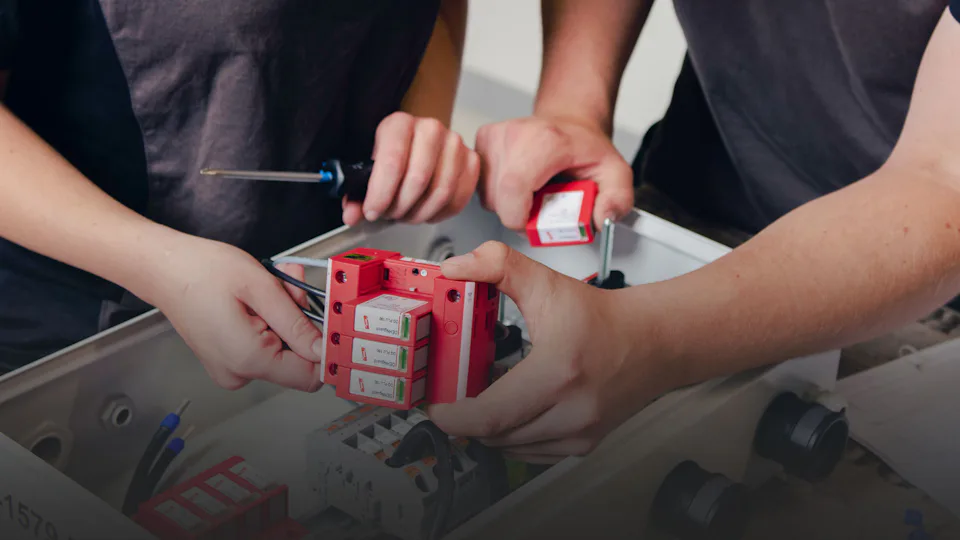 Service und Wartung Zwei Personen installieren gemeinsam rote Sicherungsrelais in einem Schaltschrank, während sie Werkzeuge verwenden, um die elektrischen Komponenten sorgfältig zu befestigen.