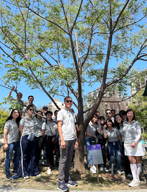 Commitment to the environment in Taiwan A group of people wearing matching gray shirts poses happily around a tall, leafy tree, enjoying a sunny day in an outdoor park setting.
