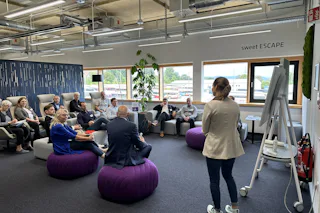 Review of the Dresden Industrial Water Day 2024 A group of people are engaged in a meeting inside a modern office space with bright natural lighting, featuring purple beanbag chairs, a whiteboard, and large windows overlooking an outdoor landscape.