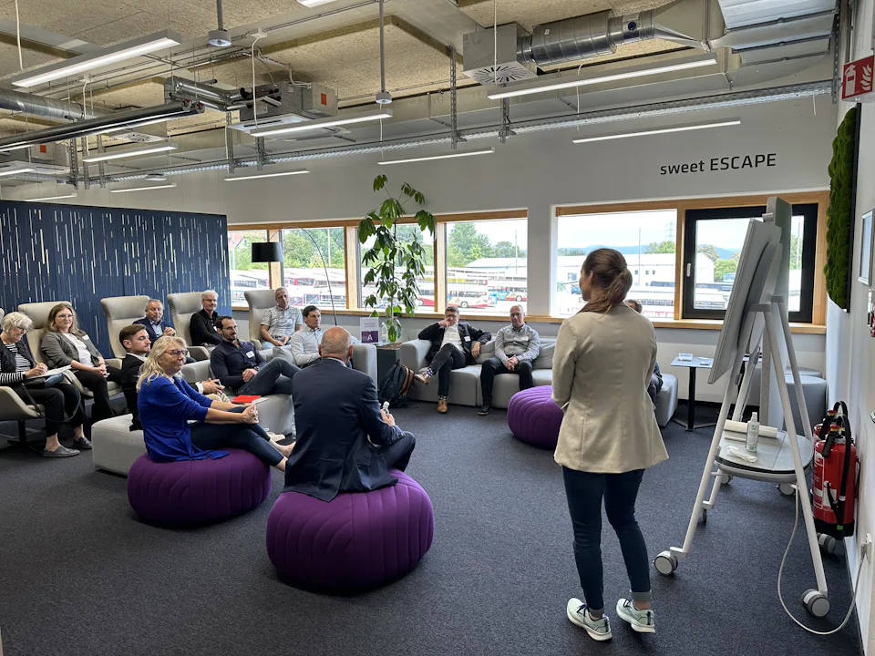 Review of the Dresden Industrial Water Day 2024 A group of people are engaged in a meeting inside a modern office space with bright natural lighting, featuring purple beanbag chairs, a whiteboard, and large windows overlooking an outdoor landscape.