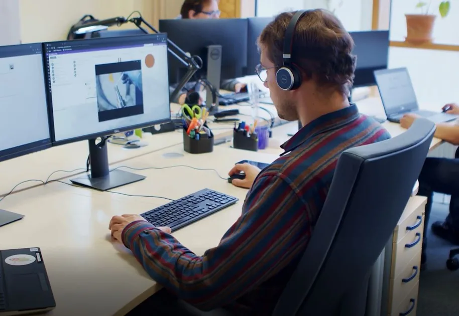 Individual Contact Person A person wearing headphones is working at a desk with three computer screens in an office; a technical video is playing on one screen.