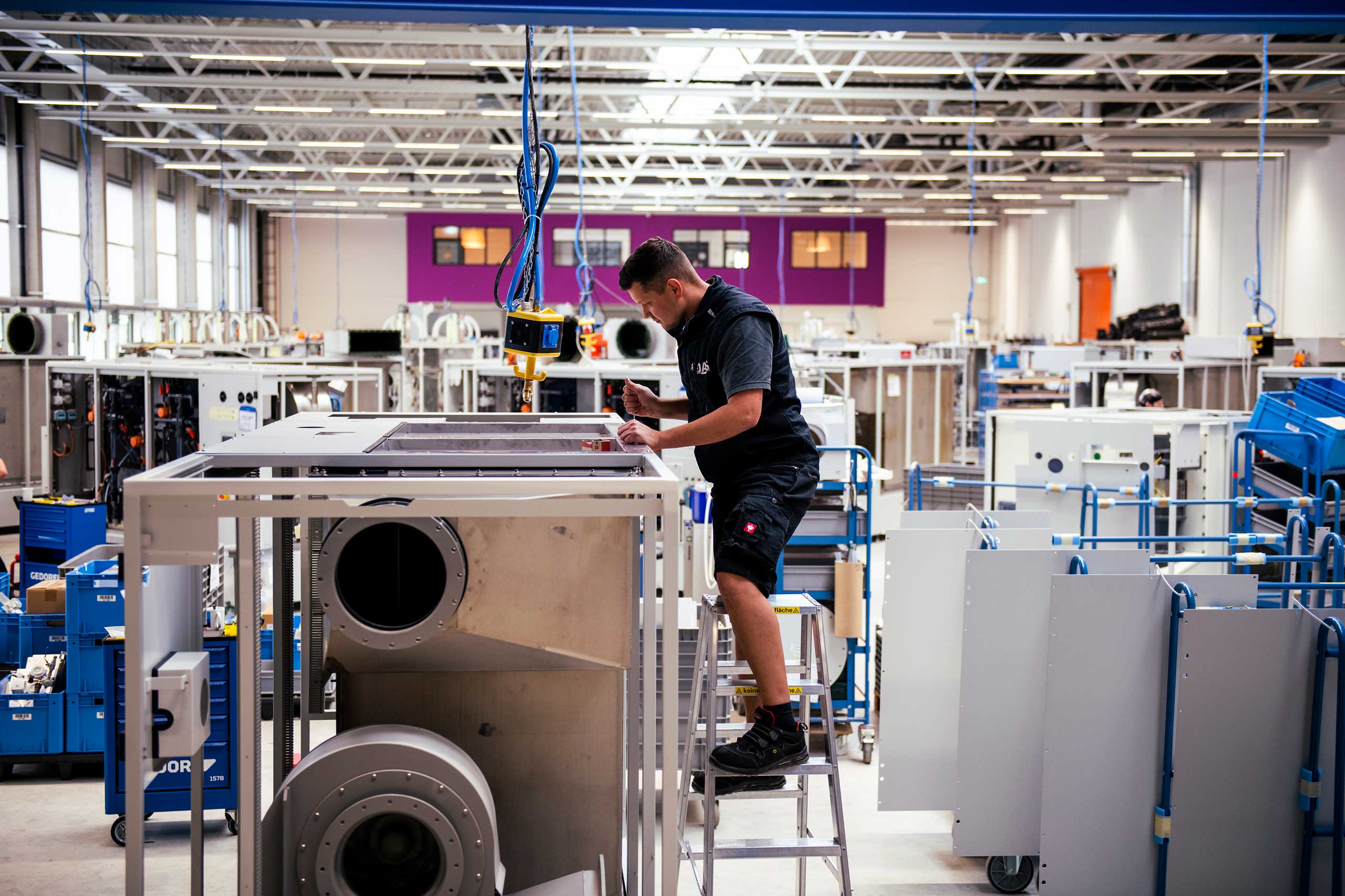 A worker stands on a ladder inspecting large industrial machinery in a spacious, well-lit manufacturing facility, surrounded by various equipment and assembly stations.