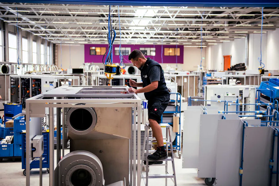 Waste Gas Treatment Manufacturing A worker stands on a ladder inspecting large industrial machinery in a spacious, well-lit manufacturing facility, surrounded by various equipment and assembly stations.