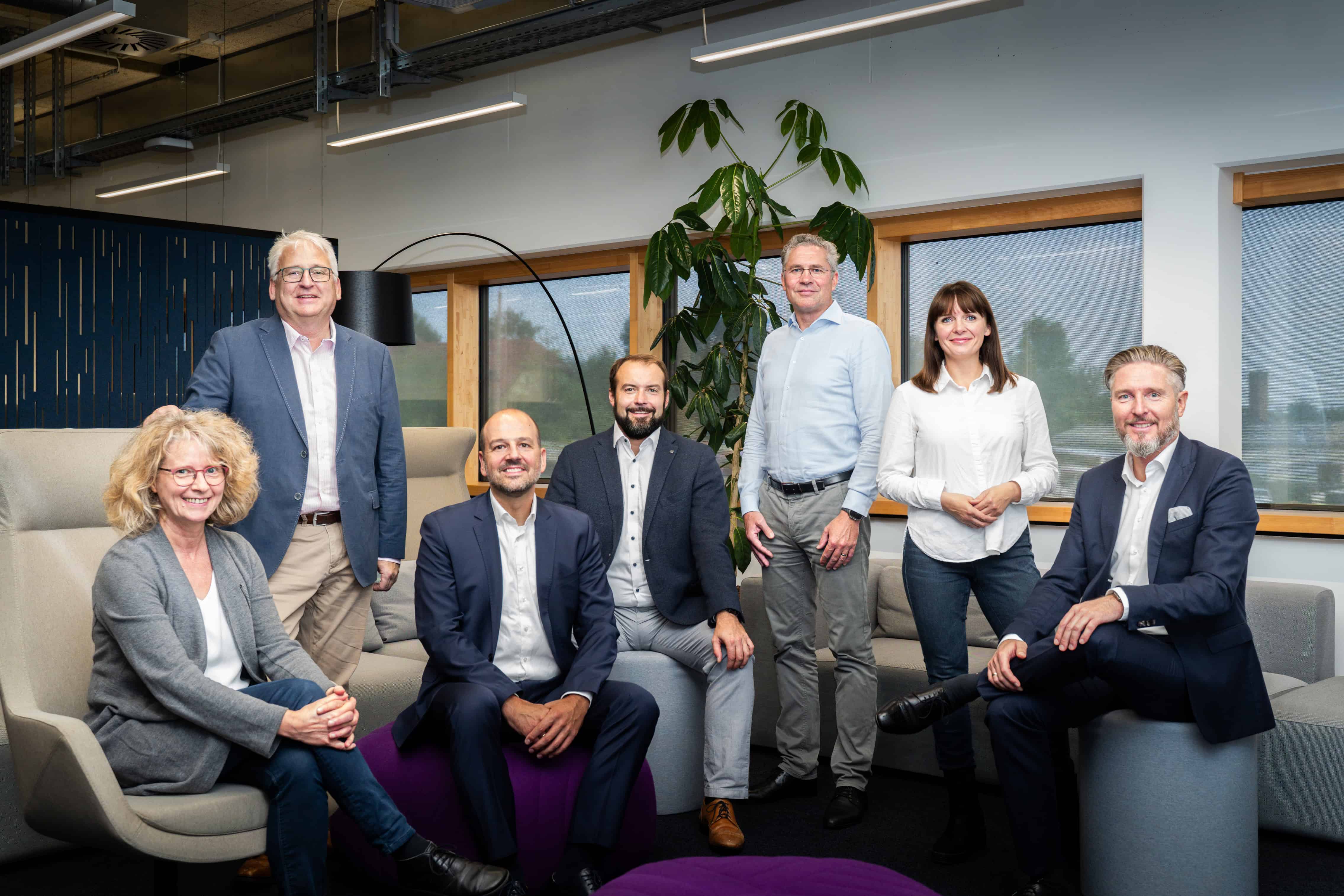 A group of seven professionals, dressed in business attire, are gathered in a modern office space with large windows and a tall potted plant in the background, seated and standing on comfortable chairs and sofas.