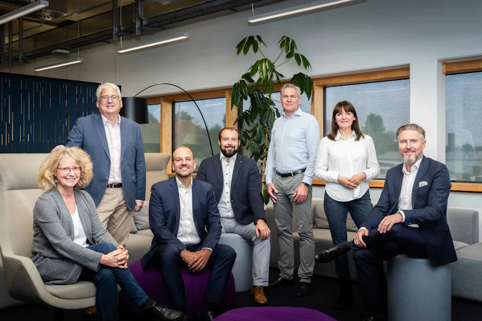DAS Management Team A group of seven professionals, dressed in business attire, are gathered in a modern office space with large windows and a tall potted plant in the background, seated and standing on comfortable chairs and sofas.