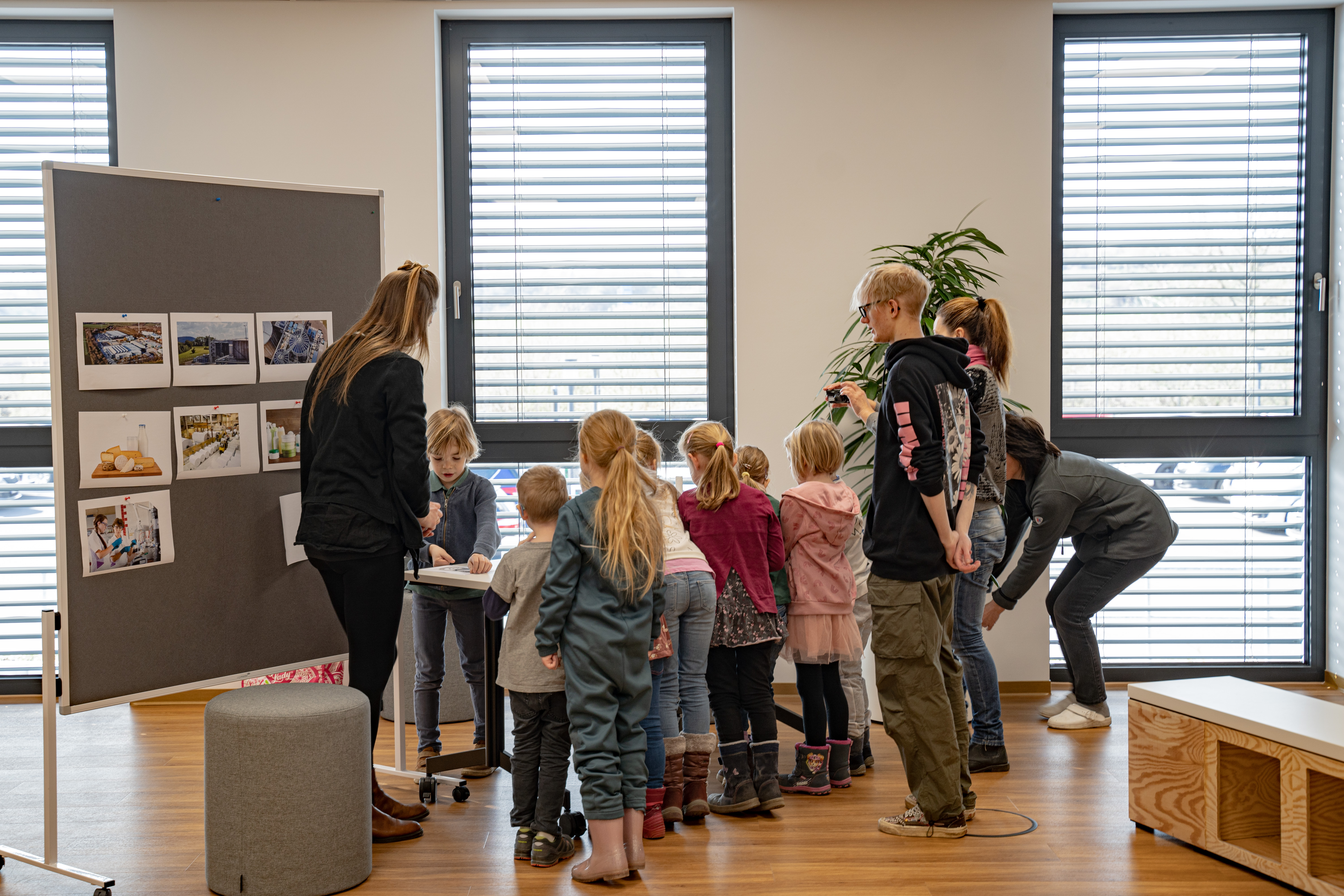 A group of children accompanied by adults attentively examines educational materials on a display board in a well-lit room with large windows, illustrating a company visit as part of a CSR educational initiative aimed at youth development and social engagement within the local community.