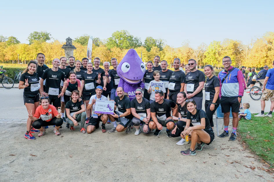 Running for a good cause: Social commitment A group of diverse individuals in athletic gear gather together in a park, alongside a purple mascot, celebrating a charity run event on a sunny day with autumn trees in the background, embodying a spirit of social engagement and community involvement.