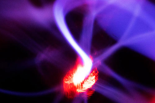 The image shows a luminous, curved plasma jet with intense violet and red colours, contrasting sharply against a dark background. A bright, glowing focal point can be seen in the centre, from which bands of light spread out dynamically.