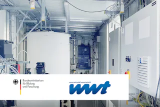 WWT-Artikel Innenansicht eines modernen Labors mit großen weißen Behältern, verschiedenen Rohren und elektrischen Schaltschränken, versehen mit den Logos des Bundesministeriums für Bildung und Forschung sowie der wwt auf einem Whiteboard.