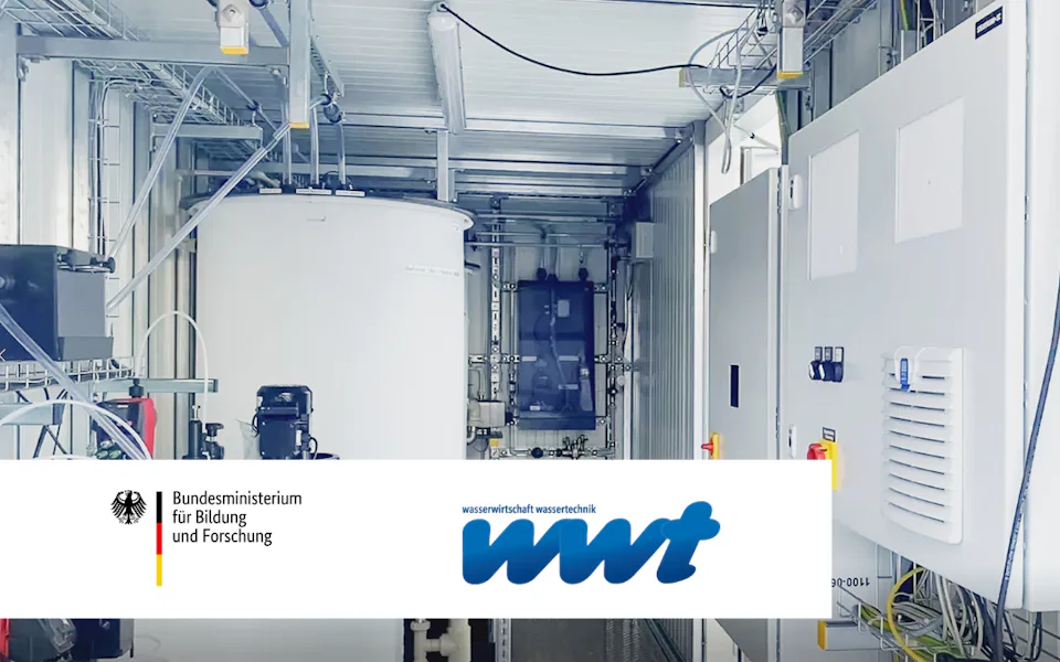 WWT-Artikel Innenansicht eines modernen Labors mit großen weißen Behältern, verschiedenen Rohren und elektrischen Schaltschränken, versehen mit den Logos des Bundesministeriums für Bildung und Forschung sowie der wwt auf einem Whiteboard.