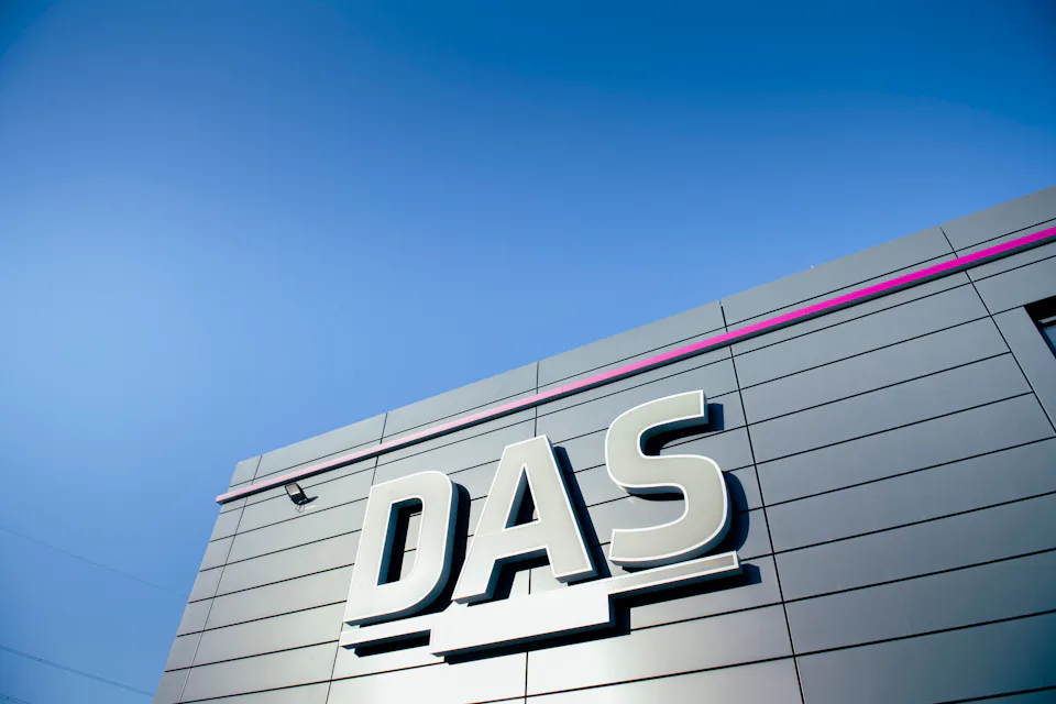 The image shows a modern industrial building facade with large white letters "DAS" prominently displayed against sleek, gray panels under a clear blue sky, emphasizing contemporary architecture and branding.