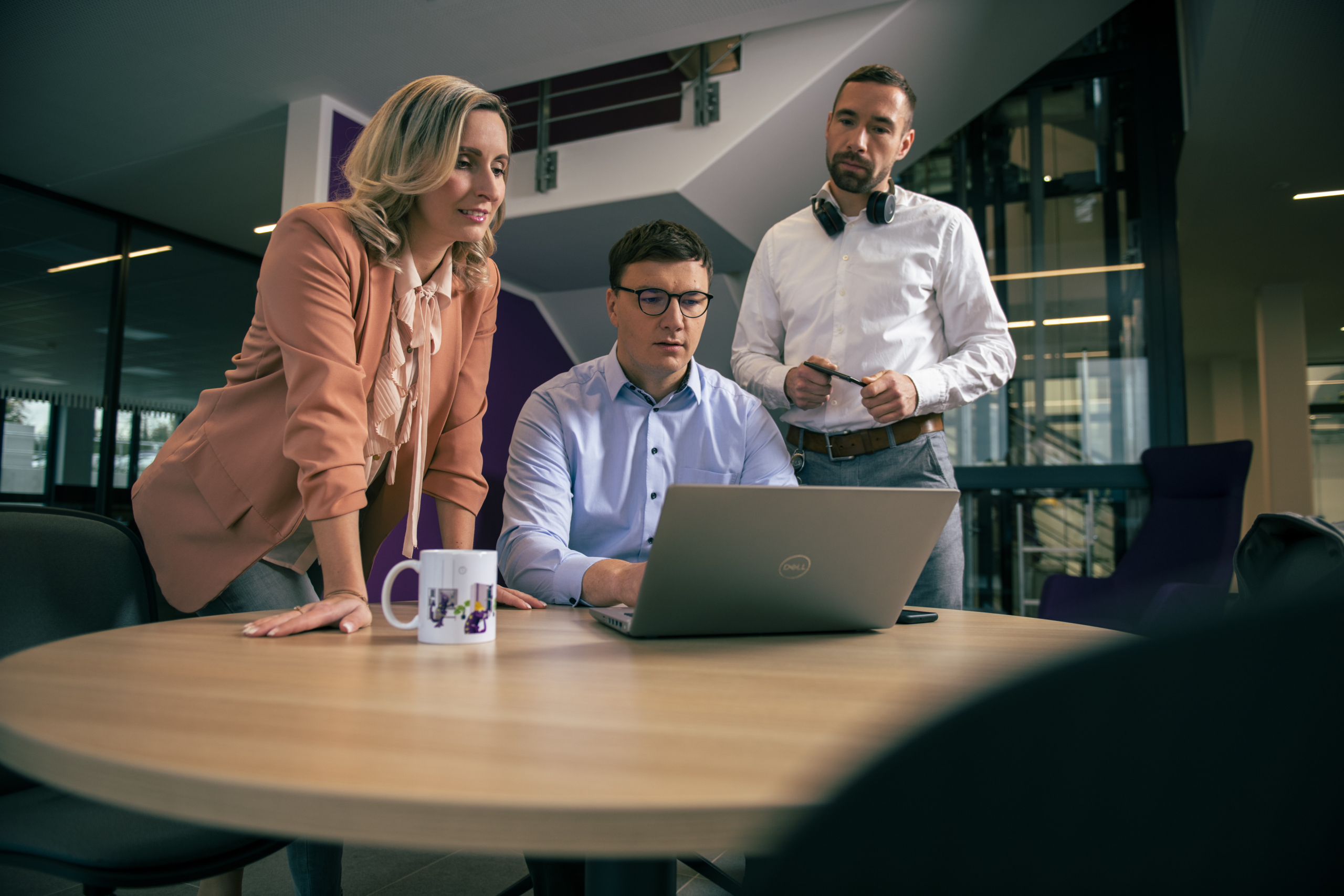 Three people collaborate around a wooden table in a modern office setting, with one person seated and focused on a laptop while the others stand attentively, surrounded by sleek furniture and contemporary architecture.