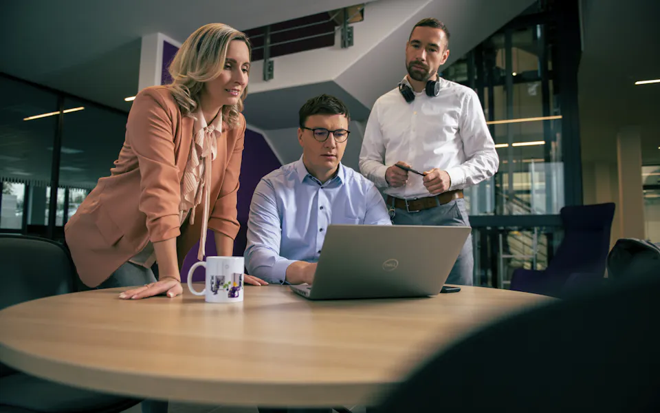 HR Team Three people collaborate around a wooden table in a modern office setting, with one person seated and focused on a laptop while the others stand attentively, surrounded by sleek furniture and contemporary architecture.