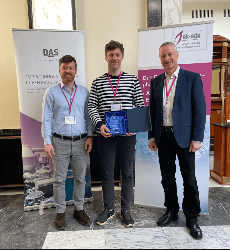 Three people stand in front of DAS and ak-adp banners on a marble floor, with one person holding a plaque, signifying an award or achievement in a formal setting.