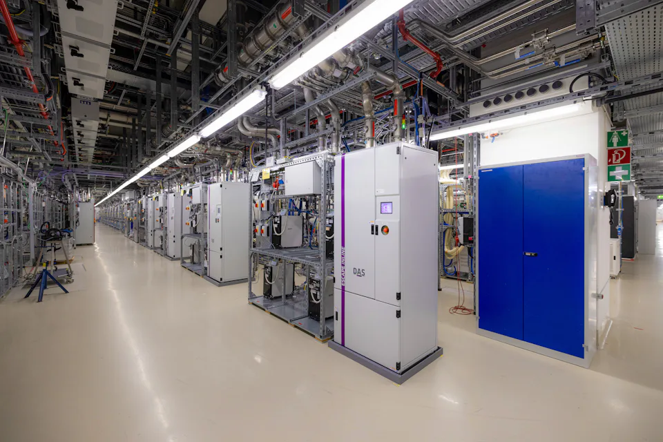 A modern industrial space houses several machines with complex piping and switch cabinet inf