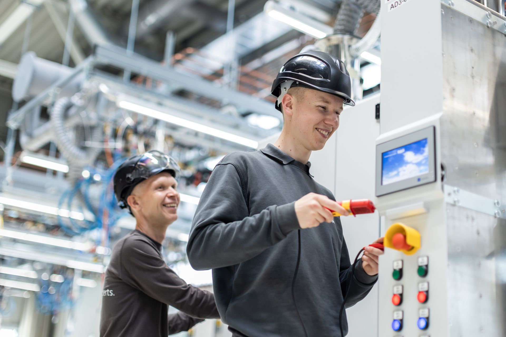 Two workers wearing black helmets and gray uniforms are smiling while interacting with control panels in a bright, modern factory setting, with visible machinery and pipes in the background.
