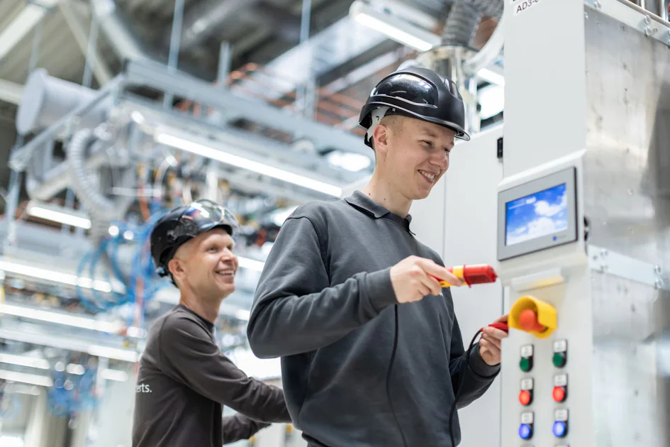 Waste Gas Treatment Two workers wearing black helmets and gray uniforms are smiling while interacting with control panels in a bright, modern factory setting, with visible machinery and pipes in the background.