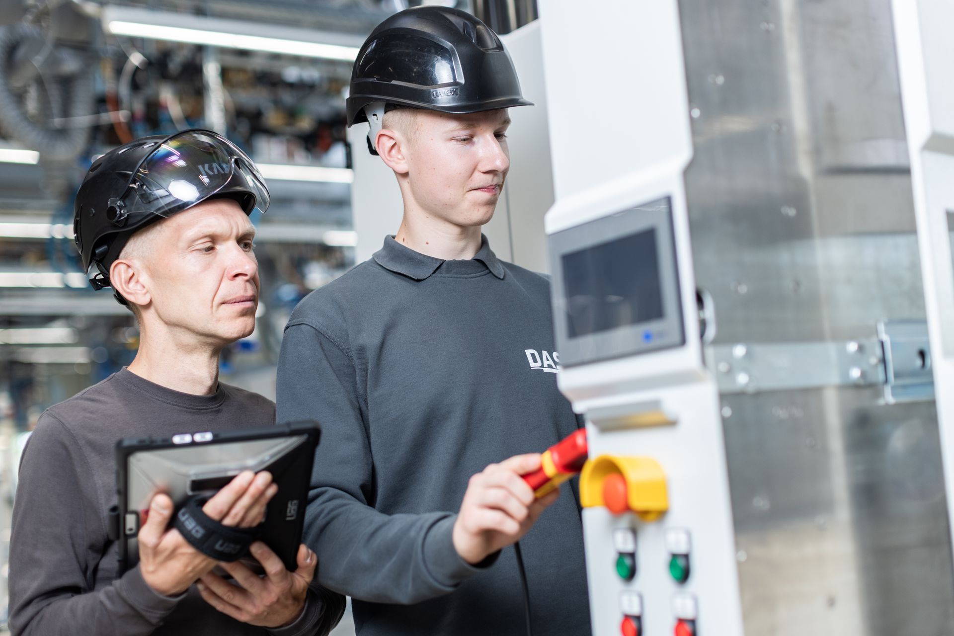 Two factory workers wearing safety helmets operate a control panel in a modern industrial setting, with one worker using a digital tablet and the other adjusting machine controls, highlighting teamwork and technology in the manufacturing industry.