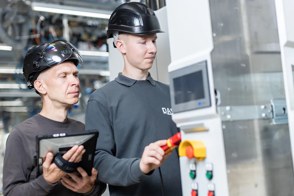 Lars Scholz Loris Gärtner Two factory workers wearing safety helmets operate a control panel in a modern industrial setting, with one worker using a digital tablet and the other adjusting machine controls, highlighting teamwork and technology in the manufacturing industry.