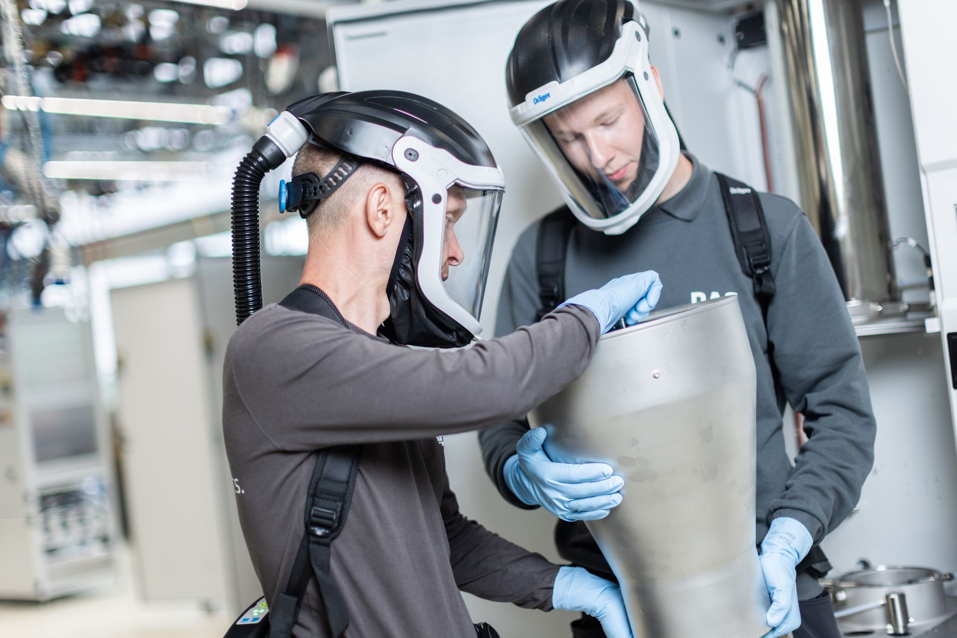 Two individuals wearing protective face shields and gloves work collaboratively with industrial equipment in a modern facility, focusing on handling a large metallic component.