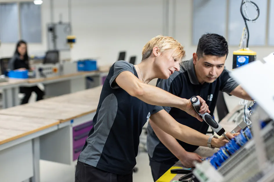 Teamwork in production: Two of our specialists work hand in hand on complex assemblies. A culture of teamwork and the highest quality standards form the basis of our efficiency and success. Two people are working collaboratively on an assembly line in a well-lit manufacturing facility, focusing intently on machinery, while another person is seen in the background at a workstation; the scene suggests teamwork and precision in a modern industrial setting.