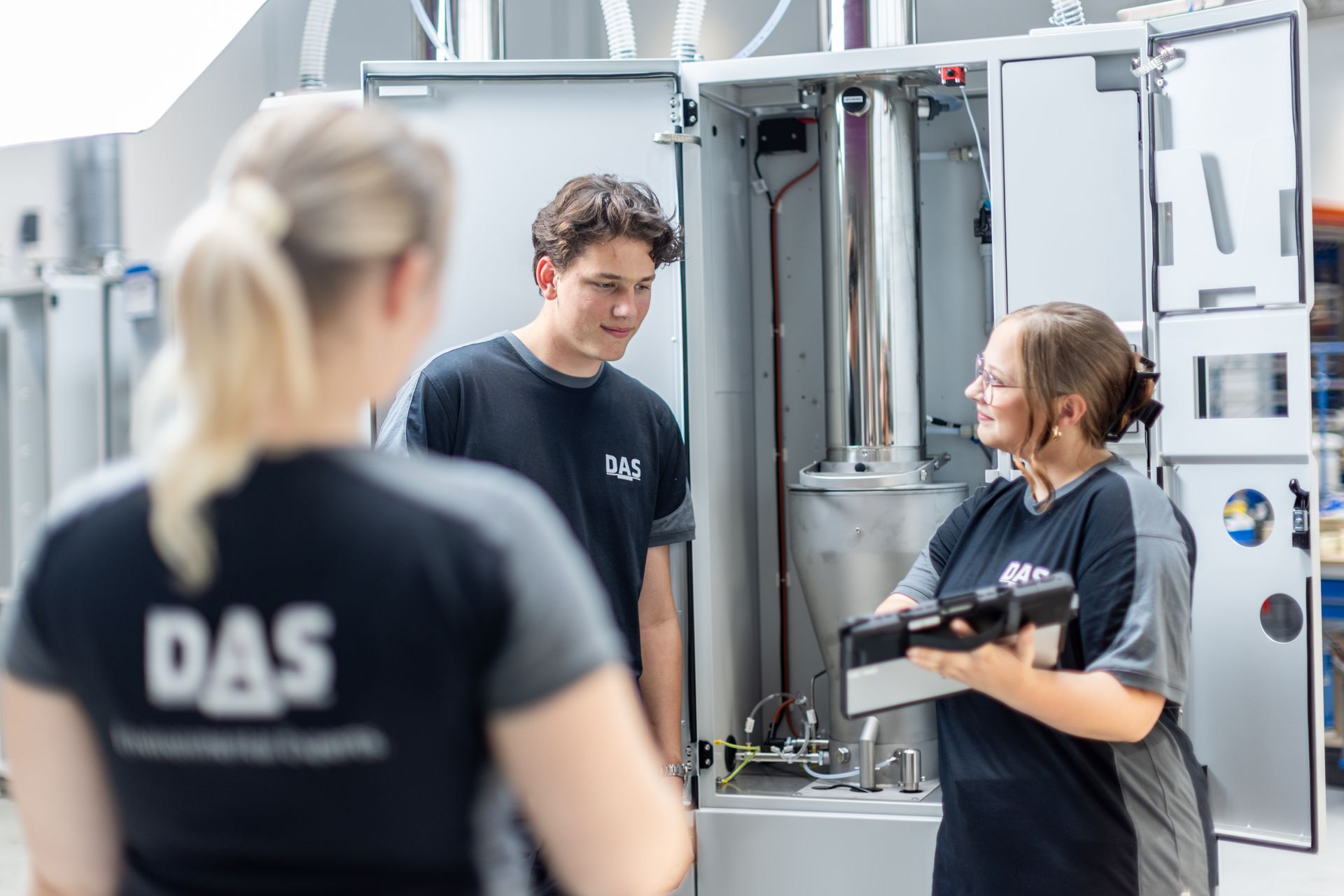 Three people in black work clothes are standing in front of an open machine, while one of them is holding a tablet and talking to a colleague.