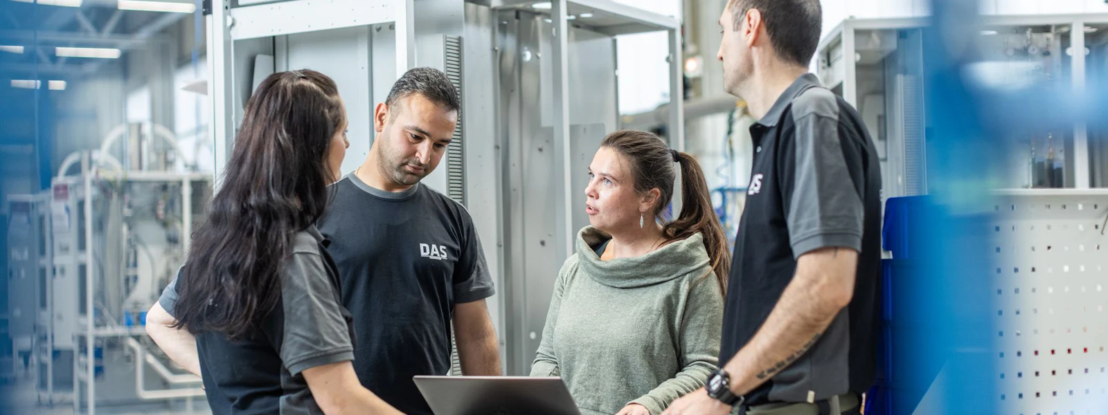 In a bright industrial facility, four workers in casual uniforms discuss logistics around a laptop on a wheeled cart, surrounded by modern manufacturing equipment.