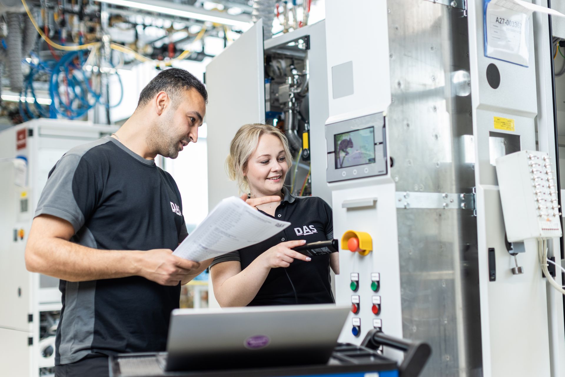 Two individuals in a factory setting engage with automated machinery and controls, discussing technical details, surrounded by industrial equipment and cables.