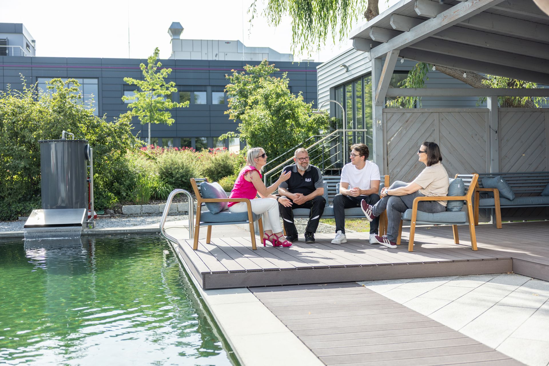 Four people sit on a wooden deck beside a serene green pond, engaging in conversation under a shaded structure, surrounded by lush greenery and modern architecture.