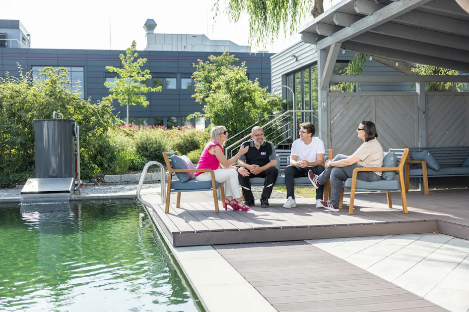 Informal meetings in green surroundings: Our outdoor area offers space for relaxed conversations, creative exchanges, and shared breaks in the natural biotope. Because a pleasant working atmosphere is important to us. Four people sit on a wooden deck beside a serene green pond, engaging in conversation under a shaded structure, surrounded by lush greenery and modern architecture.