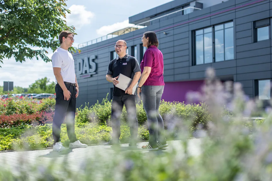 Our culture of equal opportunity Three people engage in conversation outside a modern office building with a logo, surrounded by lush greenery and flowering shrubs under a clear blue sky.